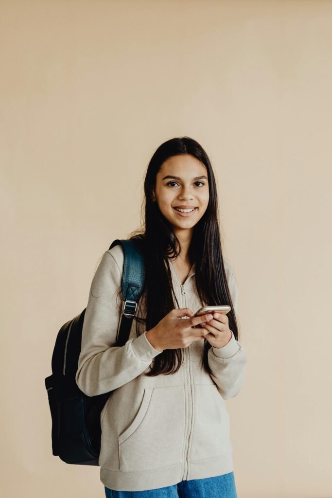 Teenage girl smiling while using smartphone, wearing a backpack, against a neutral background.